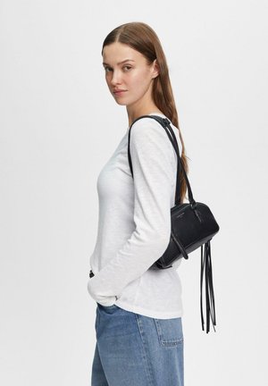 Kobieta w białej bluzce z długim rękawem i niebieskich dżinsach niesie małą czarną torebkę na ramię z frędzlami na jednolitym białym tle.