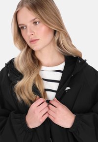 Fekete, cipzáras kapucnis dzseki, amelyen a mellrészen fehér, hímzett esernyő logó látható. Fehér és tengerészkék csíkos pulóver fölött viselve.