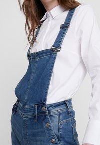 Jeansoverall med justerbara blå remmar, en framficka och sidoknappar. Bärs över en vit skjorta med krage, som har en slitna finish.