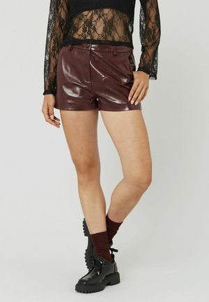 Femme portant des shorts en cuir bordeaux foncé brillant, un haut en dentelle noire transparente à manches longues, des chaussettes bordeaux foncé et des chaussures à plateforme noires.