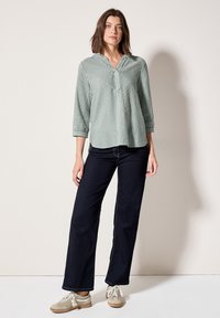 Blusa a righe verde chiaro e bianca con scollo a V e maniche a tre quarti, abbinata a jeans ampi blu scuro e scarpe beige.