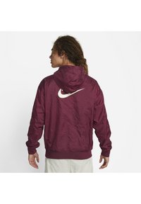 Bordo Nike jakna s kapuco iz teksturiranega materiala, z belim swoosh logotipom na hrbtu, elastičnimi manšetami in kapuco na vrvico.