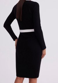 Femme portant une robe noire texturée ajustée à manches longues avec une ceinture blanche, vue de dos sur un arrière-plan clair uni.