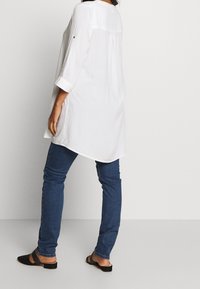 Bílá tunika s dlouhým rukávem a zakřiveným lemem, se zadečkem s přehybem, doplněná modrými skinny džíny a černými plochými sandály. Hladká textura látky.