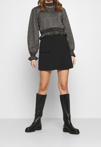 Kvinna i en metallgrå långärmad blus, svart minikjol med ficklock och svarta knästövlar, stående mot en vit bakgrund.