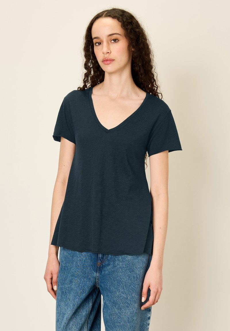 Temno modra V-neck majica iz mehkega materiala s kratkimi rokavi, ohlapnega kroja in manjšimi stranskimi razporki, v kombinaciji z svetlo modrimi kavbojkami.