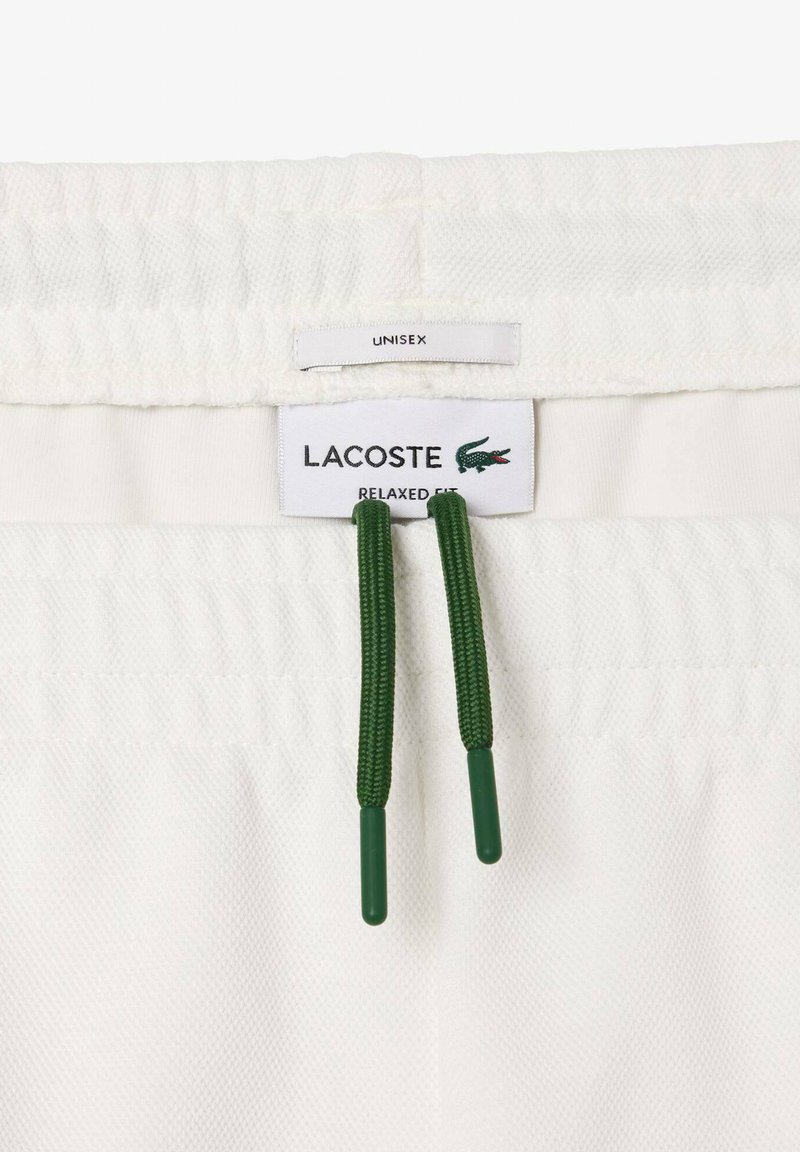 Shorts relax fit blancs Lacoste avec une taille dotée de cordons de serrage verts et un label logo Lacoste visible, ainsi qu'une étiquette unisexe.