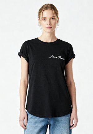 Jeune femme aux cheveux blonds porte un T-shirt noir à manches retroussées avec le texte « Mira Paris » et un jean bleu clair sur fond blanc.