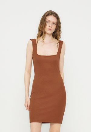 Junge Frau mit welligem Haar, die ein ärmelloses, figurbetontes braunes Kleid mit breiten Trägern trägt, steht vor einem schlichten hellen Hintergrund.