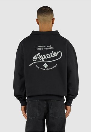 Mann von hinten gesehen, trägt ein schwarzes Sweatshirt mit weißem Text "time flies by - enjoy it, MOMENTS TO MEMORIES, Pegador" auf dem Rücken.