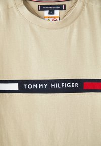 Beige puuvillane T-särk ümarate kaelustega, millel on horisontaalne must riba valgete ja punaste detailidega ning "TOMMY HILFIGER" valges tekstis.
