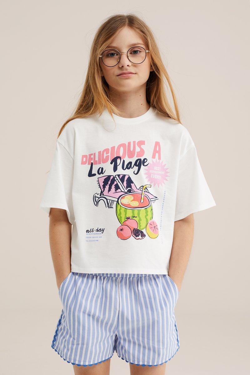 Jeune personne aux longs cheveux et lunettes portant un t-shirt blanc à motif plage et un short rayé bleu et blanc avec des bords festonnés.