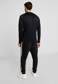 Camisa deportiva de manga larga negra con textura suave, combinada con pantalones de chándal negros con rayas blancas en los laterales y zapatillas blancas.