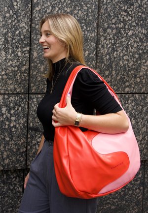 Lächelnde Frau mit blondem Haar trägt eine große rote und pinke Ledertasche über der Schulter, trägt ein schwarzes Oberteil und graue Hose vor einer Steinwand.