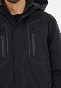 Svart, vattentät jacka med dragkedja fram och två vertikala bröstfickor. Har grå detaljer och huva. Slät textur, modern design.