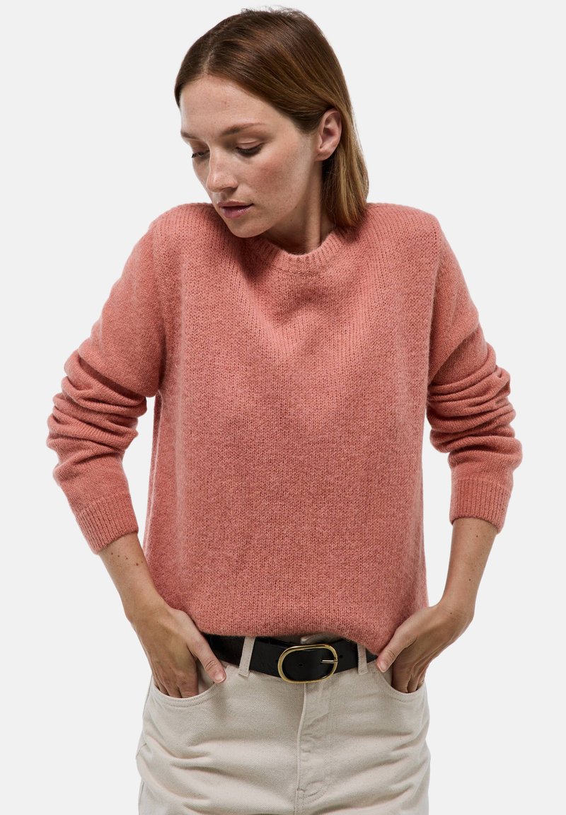 Pull en tricot rose avec un col rond, des épaules tombantes et des manches à revers, associé à un pantalon crème et une ceinture noire.