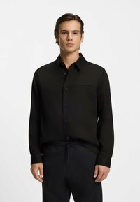 Chemise noire à manches longues avec col pointu, poche avant et texture lisse, associée à un pantalon foncé à motifs.