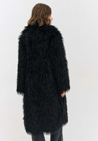 Osoba se středně dlouhými vlasy nosící dlouhý černý huňatý kabát z umělé kožešiny, obrácená zády k jednobarevnému světlému pozadí.