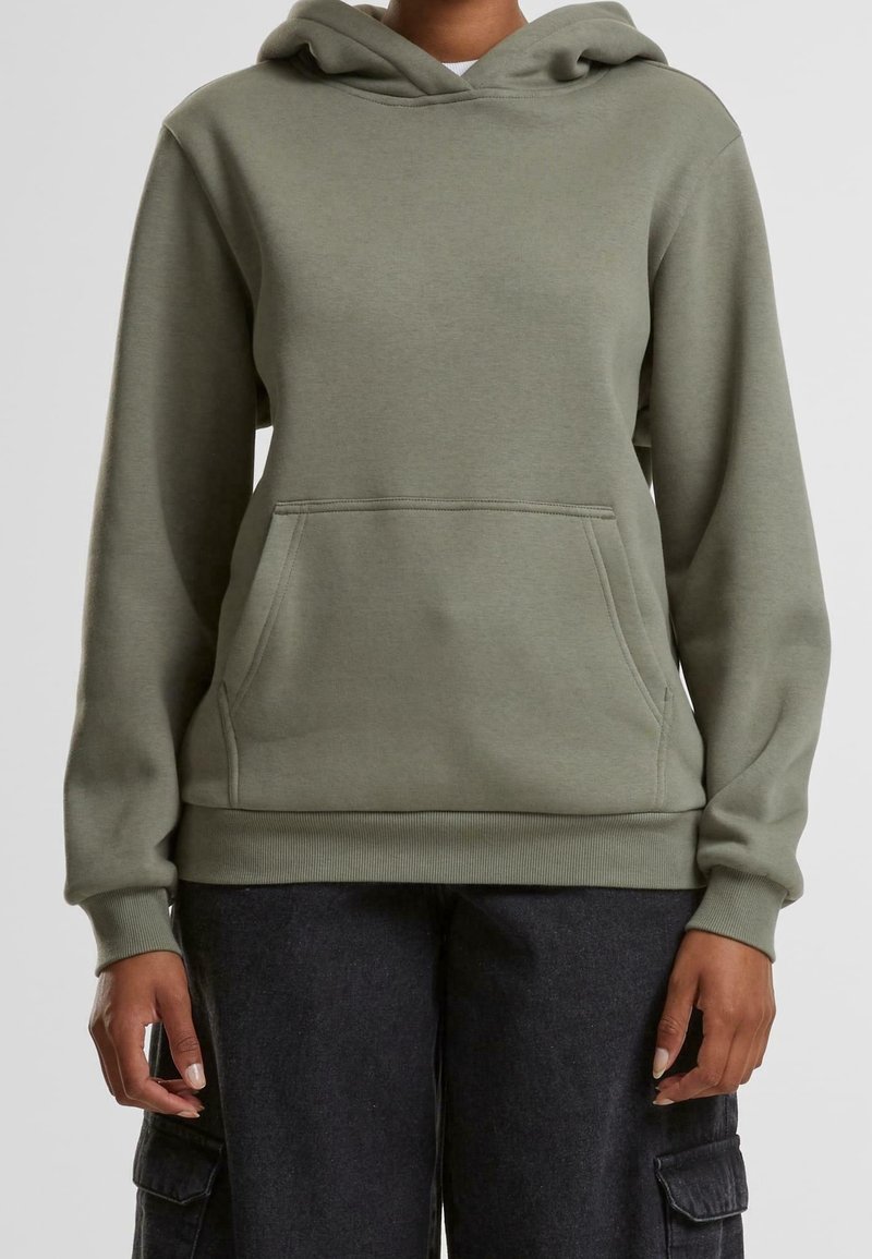 Personne portant un sweat à capuche vert olive avec une poche avant et un pantalon cargo foncé, debout devant un fond uni.