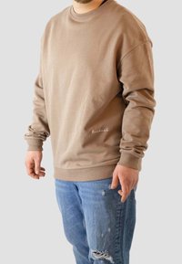 Hombre vistiendo un sudadera de cuello redondo beis y jeans azules rasgados frente a un fondo claro y liso.