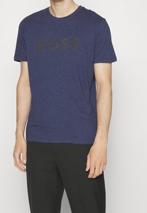 Mann in einem blauen Kurzarm-T-Shirt mit dem Schriftzug "BOSS" auf der Brust und schwarzen Hosen, stehend vor einem schlichten weißen Hintergrund.