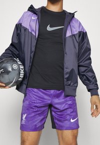 Nike windbreaker jacket in navy and purple over a black t-shirt, paired with purple striped shorts, holding a black soccer ball.