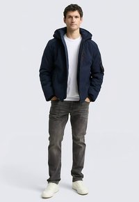 Tmavomodrá prešívaná bunda s kapucňou, vreckami so zipsom a rebrovanými manžetami, kombinovaná s bielym tričkom, sivými džínsami a bielymi teniskami.