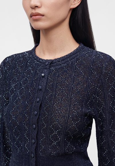 Femme portant un cardigan bleu foncé en tricot avec des fils scintillants et de petits boutons, visible des épaules jusqu'au milieu du torse.