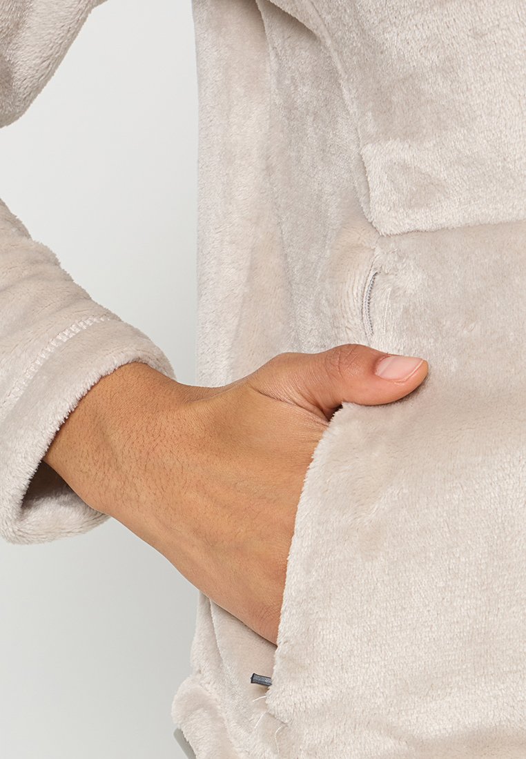 Helles beiges Fleece-Jacke mit Reißverschluss, vorderer Tasche und strukturiertem Stoff. Auffällige Details sind die genähten Bündchen und ein subtiler Glanz.