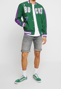 Chaqueta bomber verde y morada con letras "BUCKS", combinada con pantalones cortos grises enrollados y zapatillas verdes, con cordones y calcetines blancos.