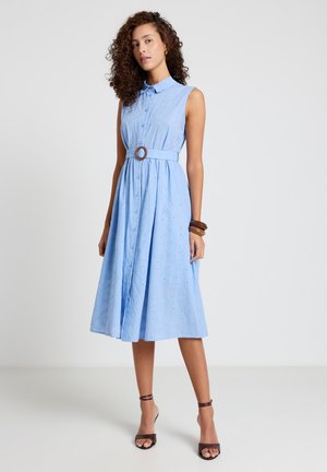 Donna che indossa un vestito midi azzurro chiaro senza maniche, con bottoni davanti, colletto e cintura, abbinato a tacchi marroni e braccialetti di legno.