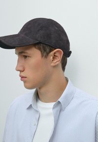 Jonge man in een zwarte baseballcap, witte t-shirt en een blauwgestreept overhemd, staat naar links gekeerd tegen een effen achtergrond.