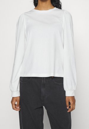 Hvid langærmet skjorte lavet af blødt stof, med rund halsudskæring og sammensyet skulderdetalje. Kombineret med mørke jeans.