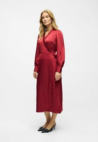 Robe enveloppante en satin rouge avec des manches longues, un col à volants et un détail de taille nouée, complétée par des escarpins noirs à bride arrière.