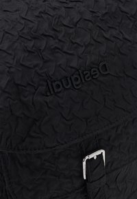 Czarny plecak o fakturze z pikowanym wzorem, z metalową klamrą w kolorze srebrnym i wytłoczonym logo marki. Wykonany z trwałego materiału nylonowego.