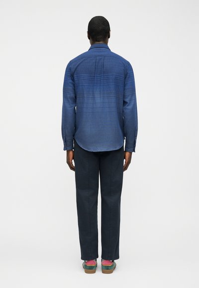 Άνδρας που στέκεται με την πλάτη στραμμένη, φοράει μπλε πουκάμισο με σχέδια και μακριά μανίκια, σκούρο παντελόνι και πολύχρωμα παπούτσια με ροζ κάλτσες πάνω σε λευκό φόντο.