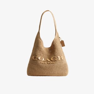Geantă de umăr țesută de culoare maro deschis, cu o singură curea și un logo mare „COACH New York” în relief pe față, cu o etichetă mică atârnată.