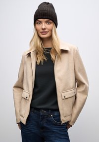 Chaqueta de mezcla de lana beige con cuello, cierre de cremallera y dos bolsillos frontales. Combinada con un suéter oscuro y jeans azules. Gorro negro de canalé.