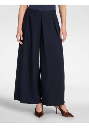 Femme portant un pantalon large taille haute bleu marine associé à des escarpins marron à bout pointu, debout devant un fond uni.
