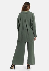 Grøn tekstureret loungewear sæt bestående af en langærmet top med krave og bredbenede bukser. Sorte højhælede sko synlige.