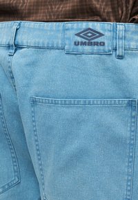 Svetlomodré rifľové nohavice s zadnými vreckami a čiernou nášivkou s logom Umbro na páse, skombinované s hnedým textúrovaným topom.