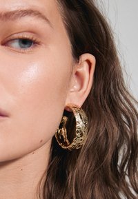 Boucles d'oreilles en or en forme de créole avec une finition texturée et martelée, de taille moyenne, positionnées sur l'oreille du modèle, complétées par des cheveux ondulés à proximité.