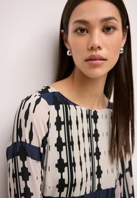 Haut en mélange de soie avec un col rond, arborant un motif géométrique noir et crème, des manches longues et une texture de tissu lisse.
