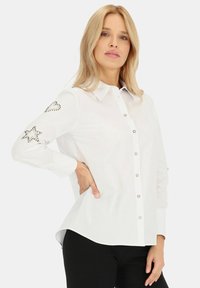 Biała koszula zapinana na guziki z długimi rękawami, zdobiona dekoracyjnymi srebrnymi ćwiekami w kształcie gwiazdek i serc na lewym rękawie. Gładki materiał, dopasowany krój.