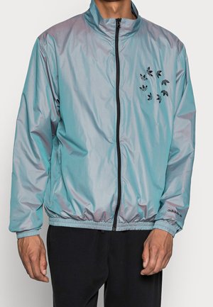 Homme portant une veste zippée Adidas bleu clair avec des motifs de logo noir sur la poitrine et les manches, associée à un pantalon noir.