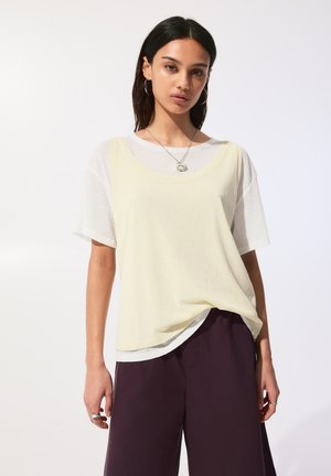 Maglietta a strati di un giallo chiaro semitrasparente, con scollatura rotonda, maniche corte e vestibilità rilassata, indossata sopra un top bianco e abbinata a pantaloni scuri.