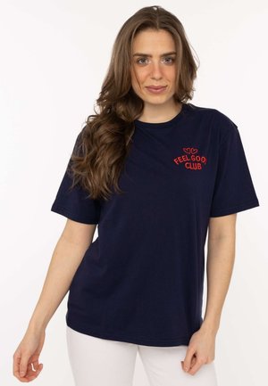 Woman with long brown hair wears navy blue t-shirt with red "Feel Good Club" text and two hearts, paired with white pants.