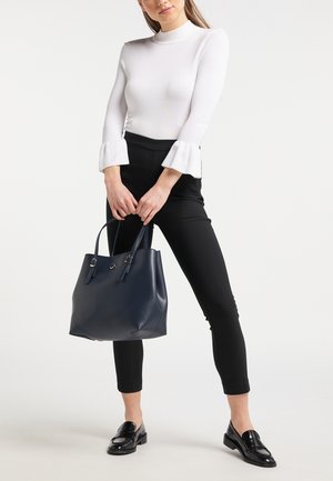 Vrouw in een witte top met lange mouwen met wijde manchetten, zwarte strakke broek, zwarte loafers, met beide handen een donkerblauwe leren handtas vasthoudend.