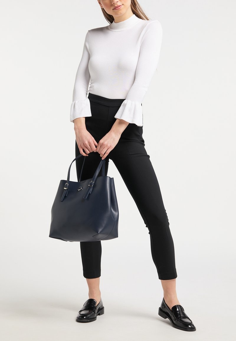 Femme portant un haut blanc à manches longues avec des poignets évasés, un pantalon noir slim, des mocassins noirs, tenant un sac à main en cuir bleu marine avec les deux mains.