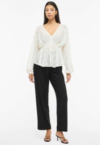 Biała koronkowa bluzka z głębokim dekoltem w kształcie litery V, długimi przezroczystymi rękawami i marszczonym pasem. Zestawiona z wysokim stanem czarnych spodni i sandałkami na paskach.